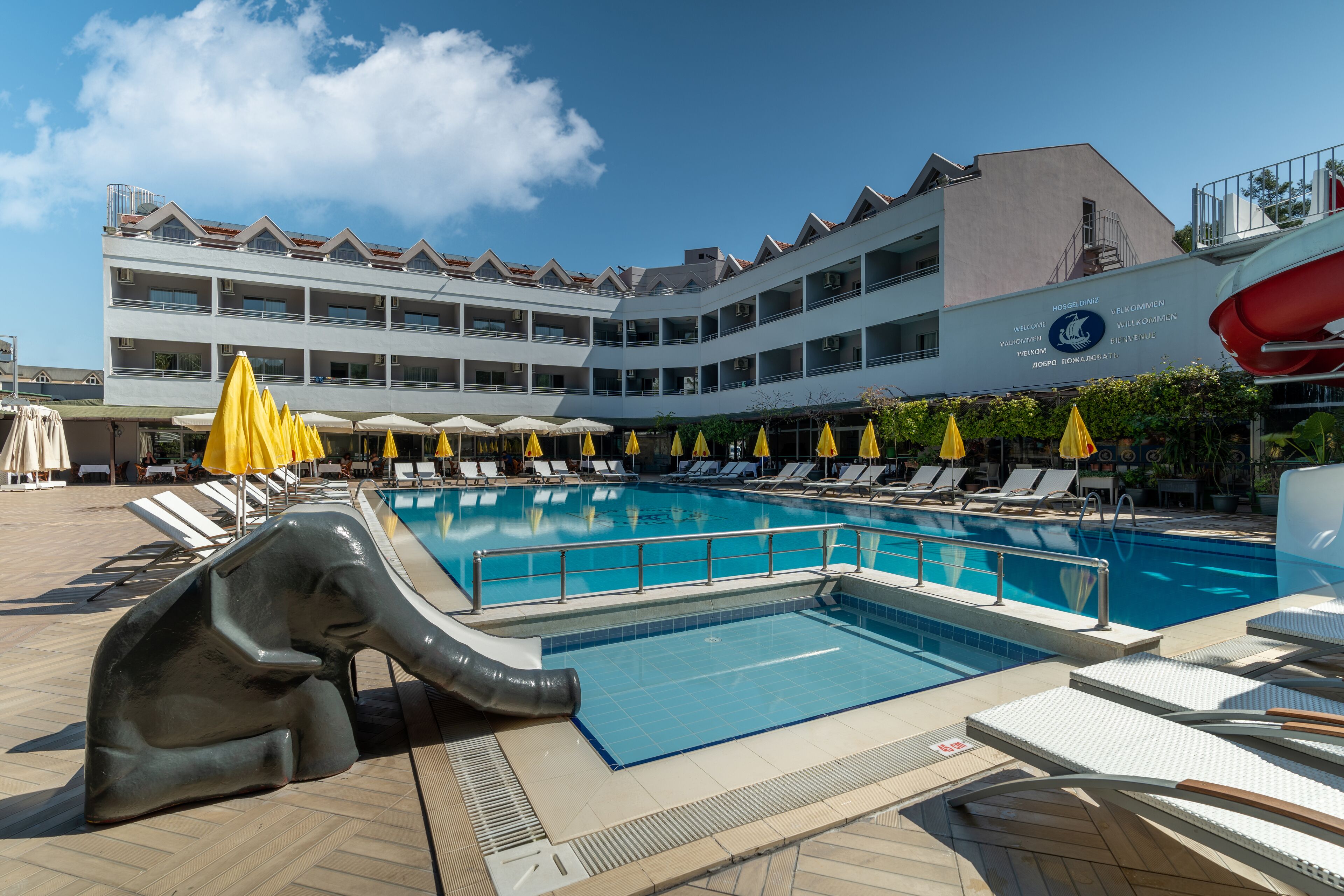 A swimming pool with lounge chairs and umbrellas, a large elephant sculpture, and a building with balconies and signage.