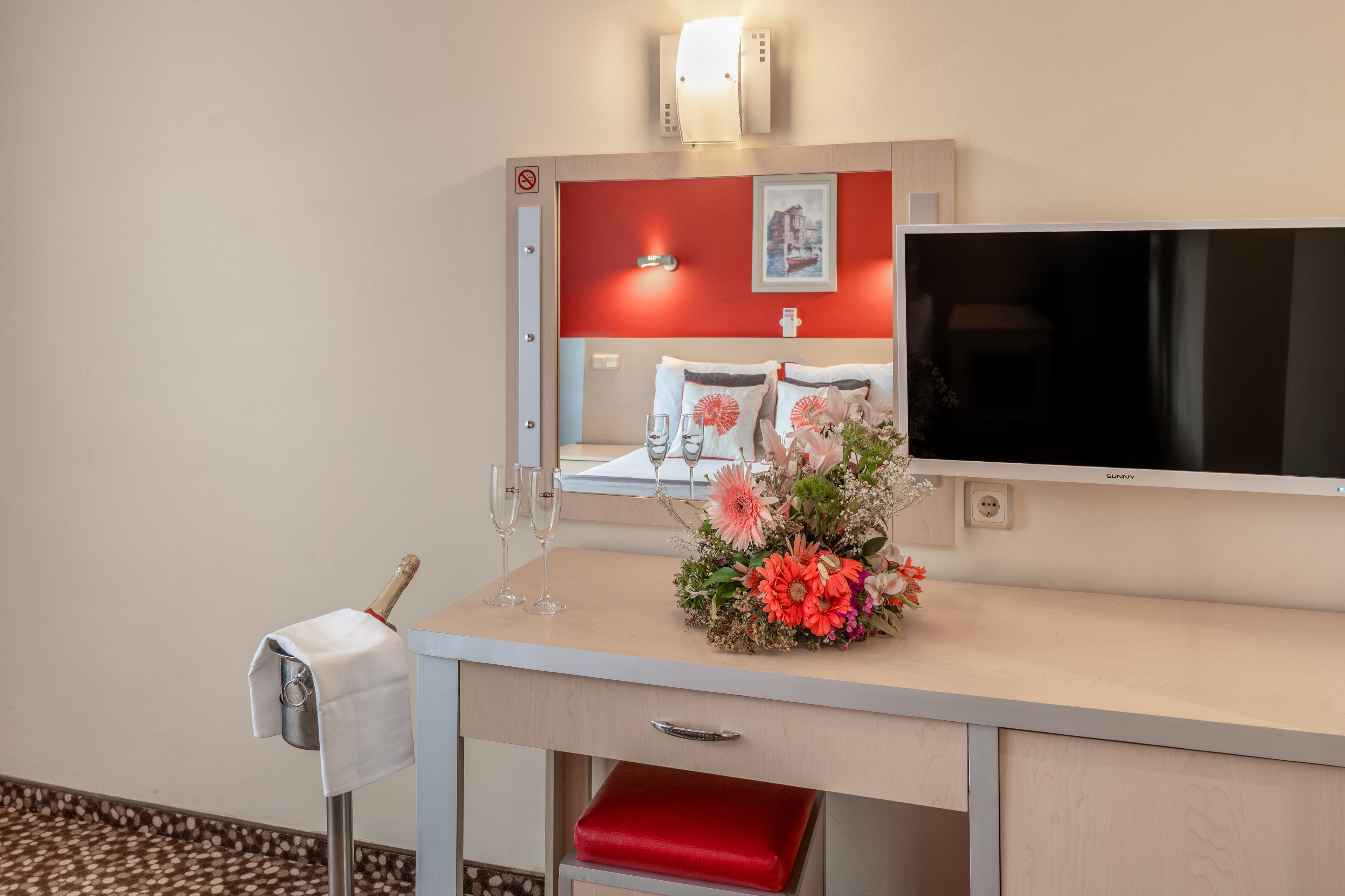 A hotel room with a bed, a desk with a mirror, a flat-screen TV, a red chair, a vase of flowers, and a minibar.