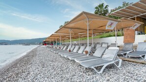 Un hotel frente al mar con filas de tumbonas bajo sombrillas, una playa de guijarros y vistas al mar y montañas distantes.