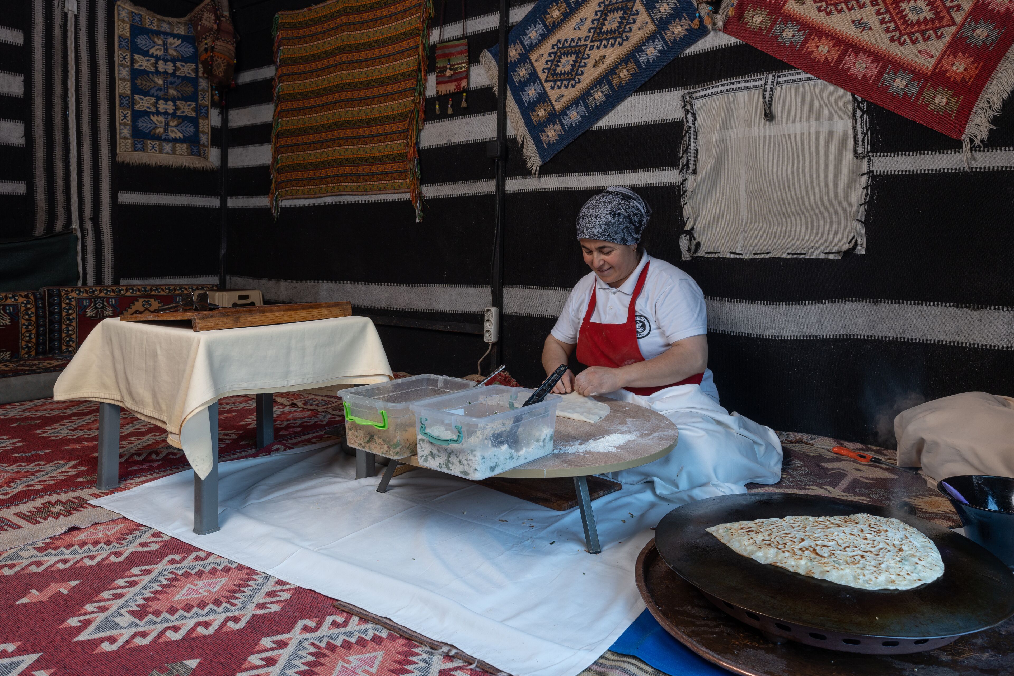 A kitchen area with a table covered by a white cloth, a large pan on the floor, and a person cooking.
