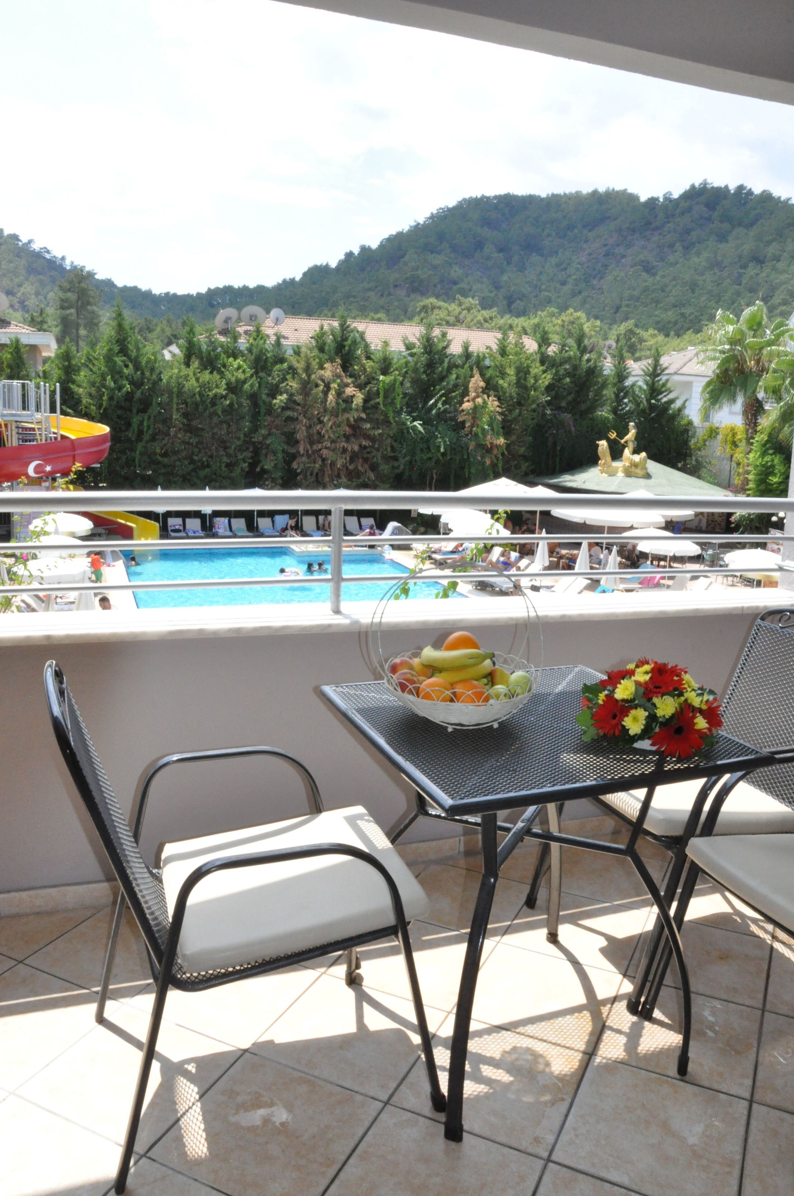 A balcony with a pool view, a table with fruit and flowers, and chairs.