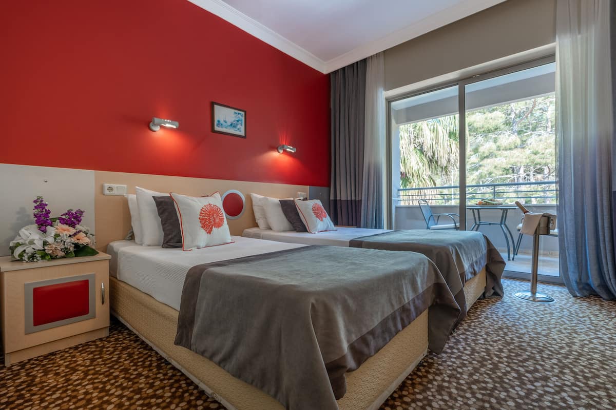 A hotel room with two beds, a red accent wall, a large window with a view, and a small table with chairs.