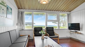 A room with a wooden ceiling, a TV on the wall, a sofa, two chairs, a table with a book and glasses, and large windows with a view of the sea.