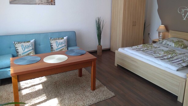 A bedroom with a bed, a wooden wardrobe, a blue sofa with beach-themed pillows, a small table with placemats, and a beach-themed wall hanging.