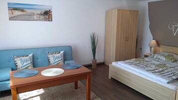 A bedroom with a bed, a wooden wardrobe, a blue sofa with beach-themed pillows, a small table with placemats, and a beach-themed wall hanging.