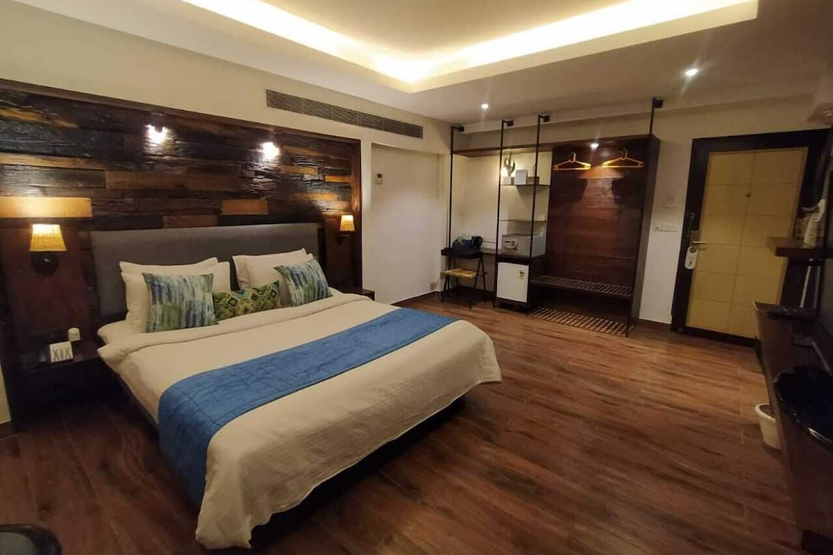A hotel room with a large bed, wooden flooring, a desk with a chair, a wardrobe, and a bathroom with a toilet and sink.