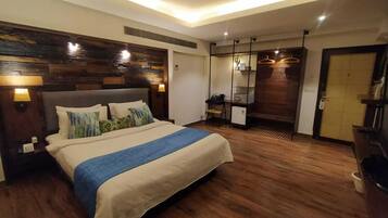 A hotel room with a large bed, wooden flooring, a desk with a chair, a wardrobe, and a bathroom with a toilet and sink.