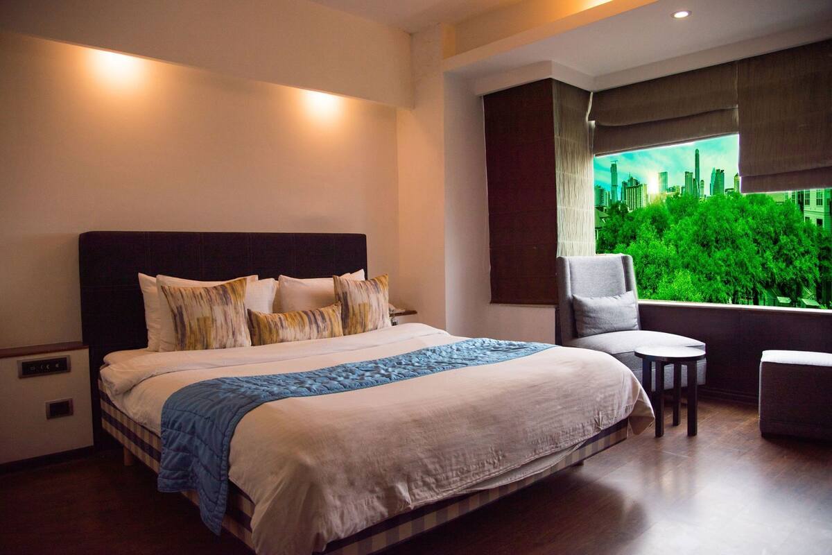 A hotel room with a large bed, a chair, a small table, and a window with a cityscape view.