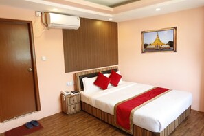 Una habitación de hotel con cabecera de madera, una cama con almohadas rojas, una mesita de noche pequeña con un teléfono y un cuadro de un templo en la pared.