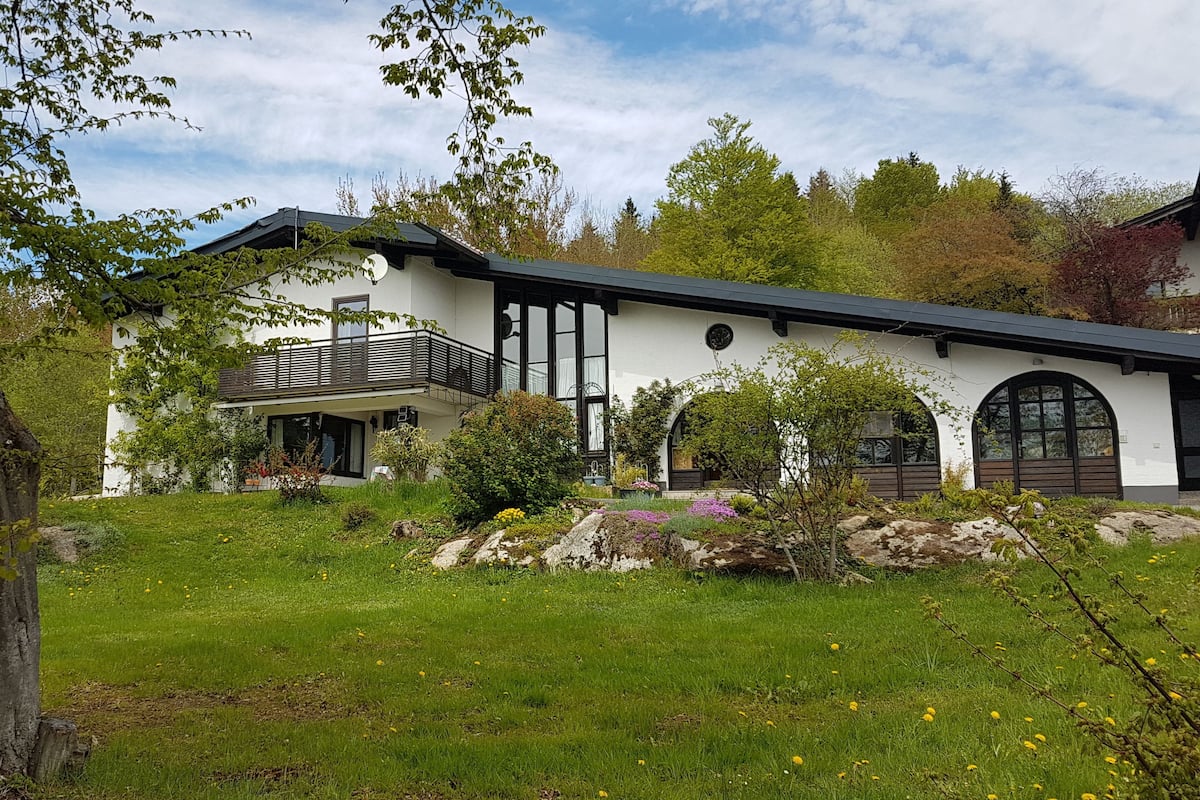 Ein modernes, zweistöckiges Haus mit Balkon, umgeben von einem gepflegten Garten mit verschiedenen Pflanzen und Blumen.