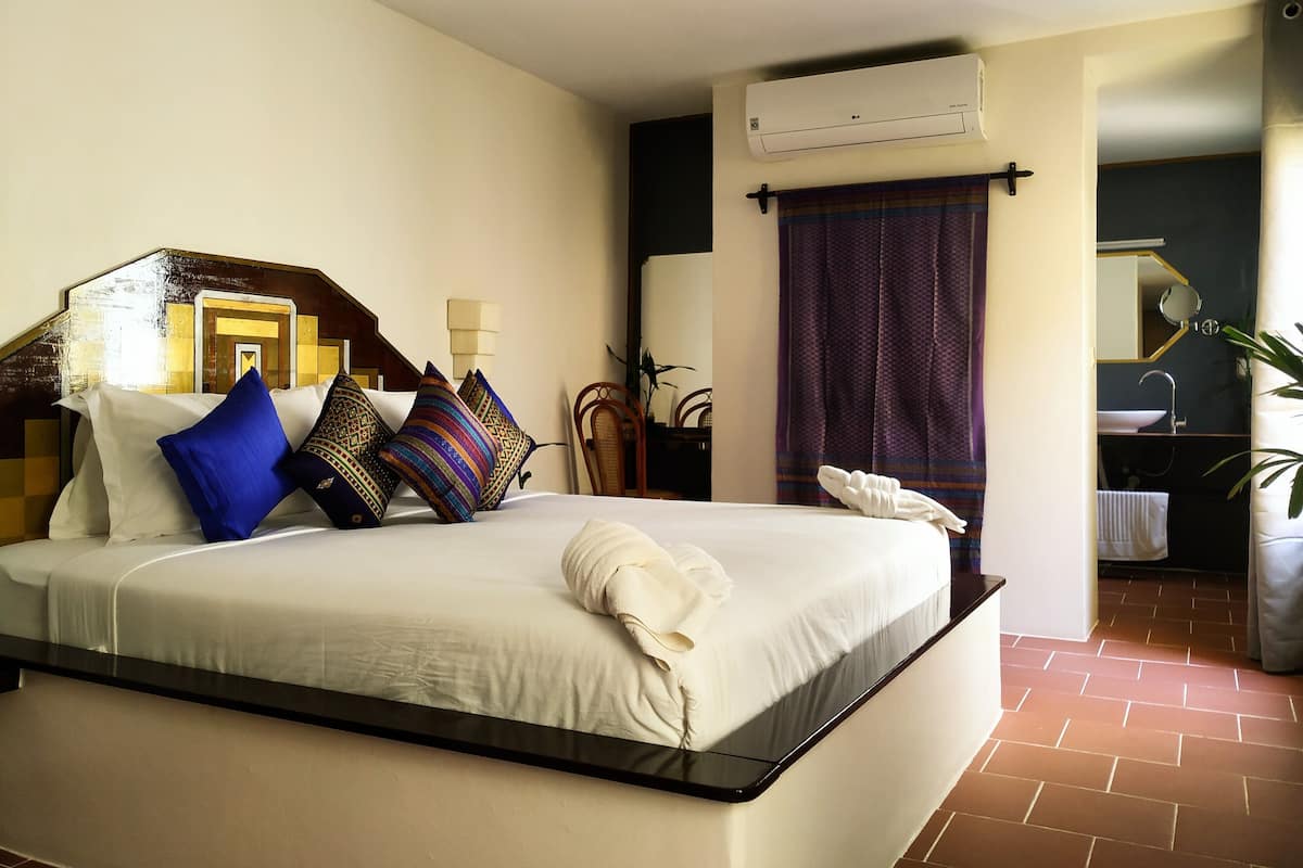 A hotel room with a large bed, a wooden headboard, a wall-mounted air conditioner, a bathroom with a sink and mirror, and a potted plant.