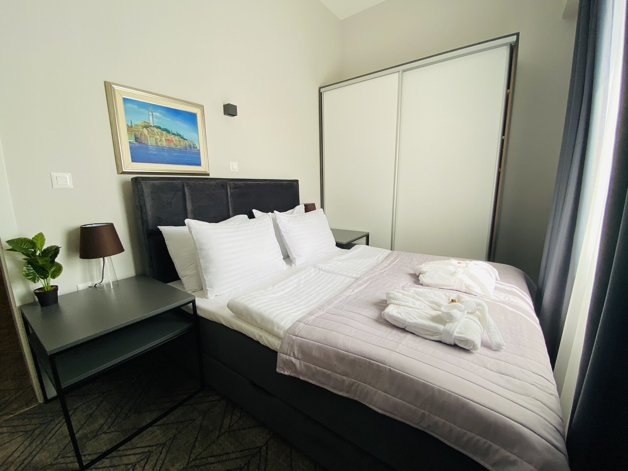 A hotel room with a bed, a nightstand, a lamp, a potted plant, and a painting on the wall.