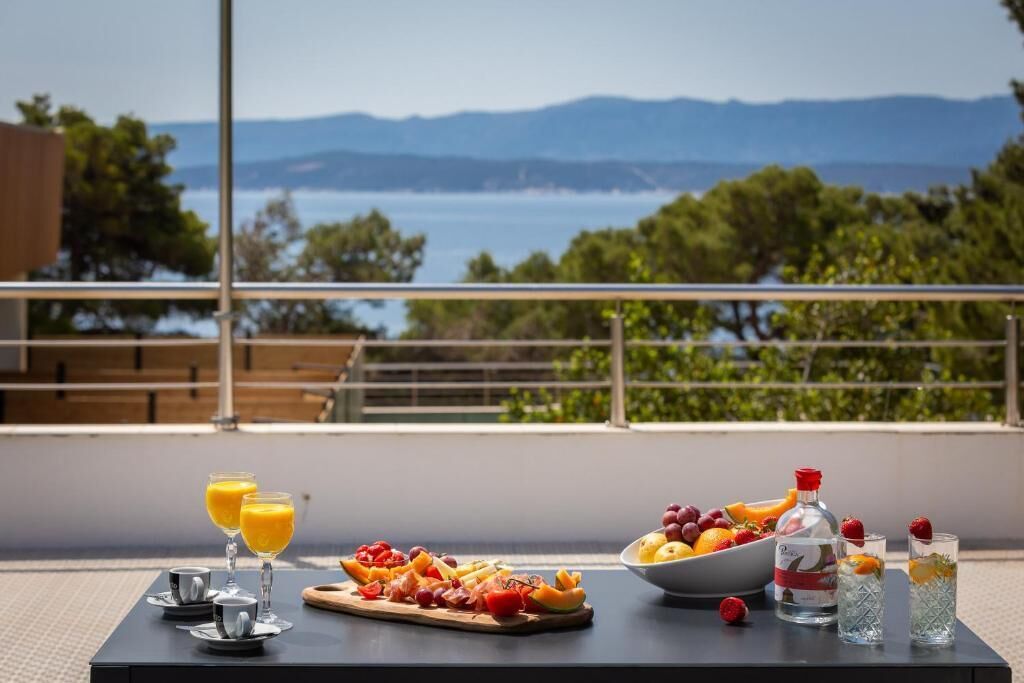Un tavolo con un vassoio di frutta, bicchieri di succo d'arancia e una bottiglia d'acqua, con vista panoramica su un lago e montagne.