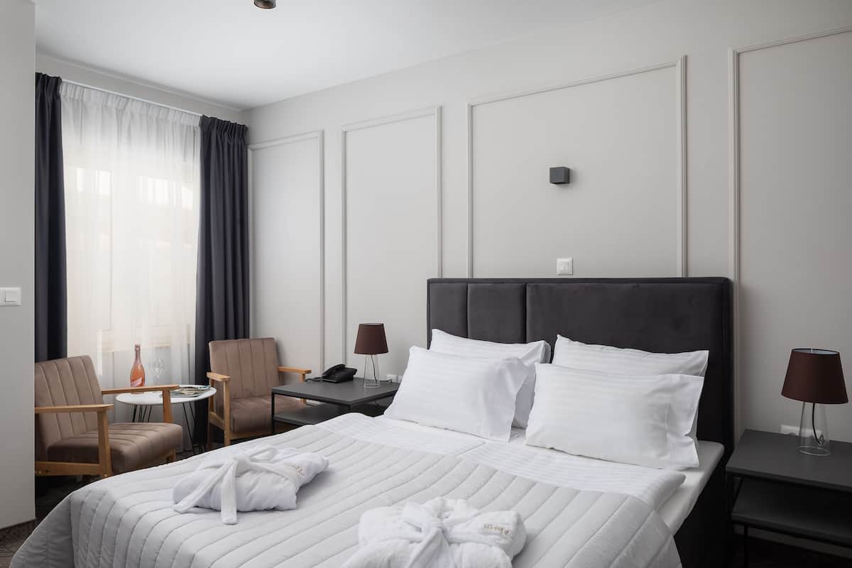 Une chambre d’hôtel avec un grand lit, deux tables de chevet, une chaise, une petite table et une fenêtre avec des rideaux.