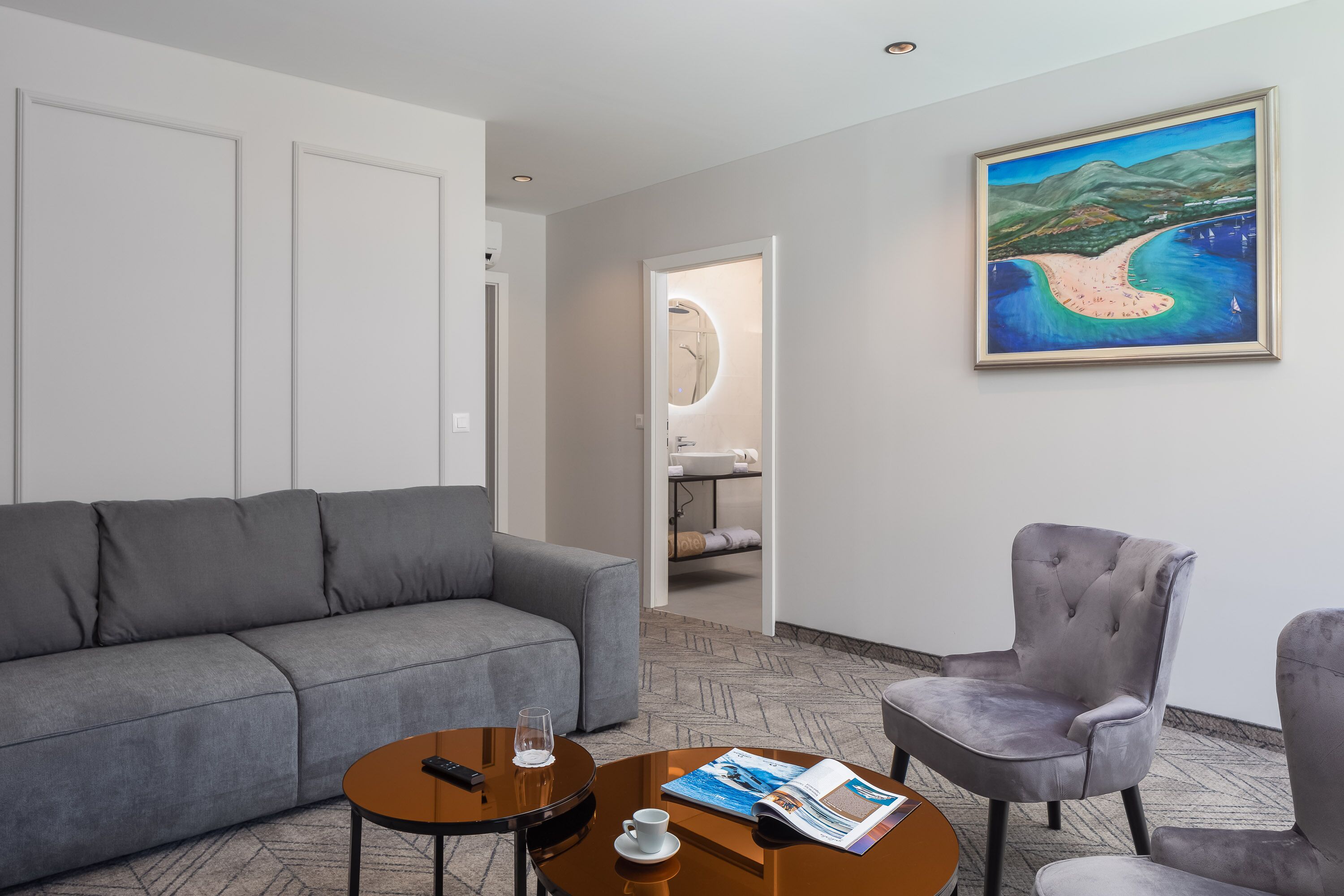 A modern hotel room with a gray sofa, a round coffee table with a remote control, a magazine, and a cup. There's a gray armchair and a painting of a landscape on the wall.