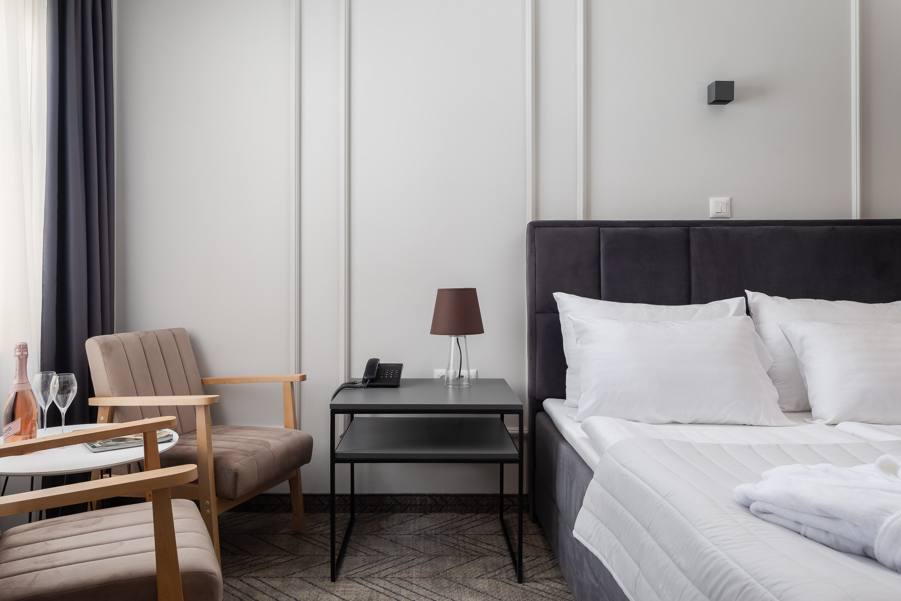 A hotel room with a bed, a chair, a small table, a lamp, and a bottle of wine.