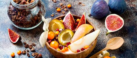 A wooden bowl filled with various fruits and nuts, a halved fig, and a wooden spoon on a textured surface.