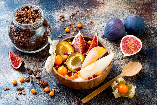 A wooden bowl filled with various fruits and nuts, a halved fig, and a wooden spoon on a textured surface.