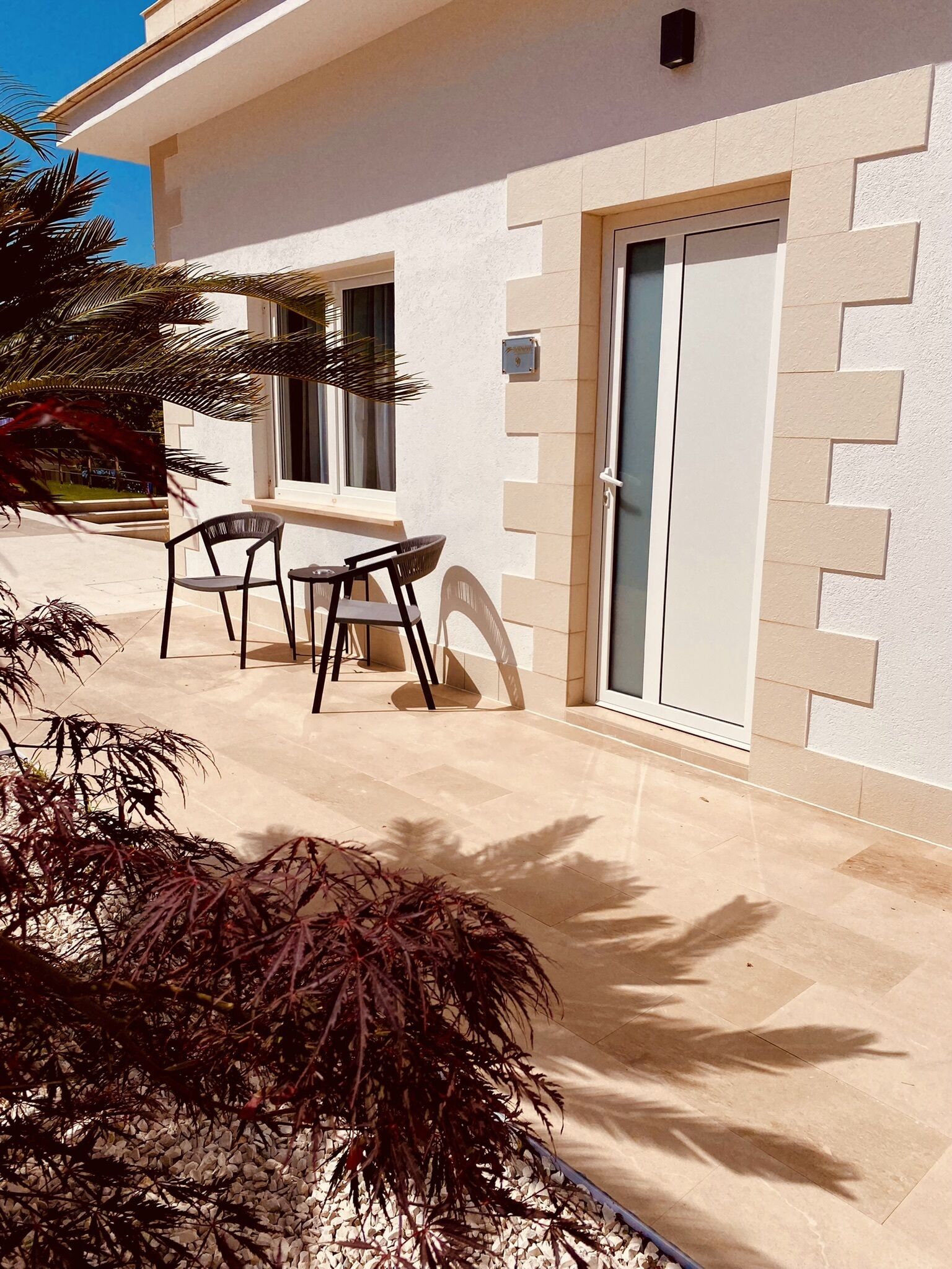 Een modern gebouw met een betegeld terras, een deur met ruitpaneel, en een kleine tafel met stoelen.