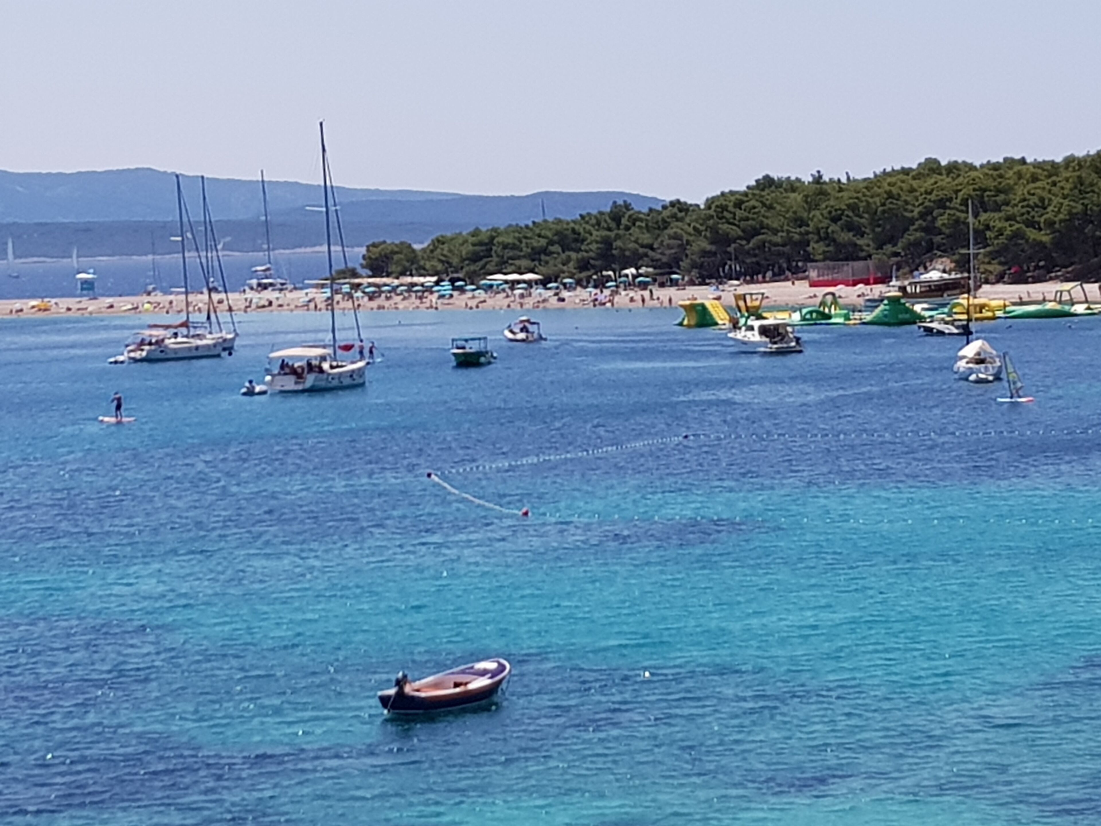 Escena costera con barcos en el agua, una playa y un área boscosa al fondo.