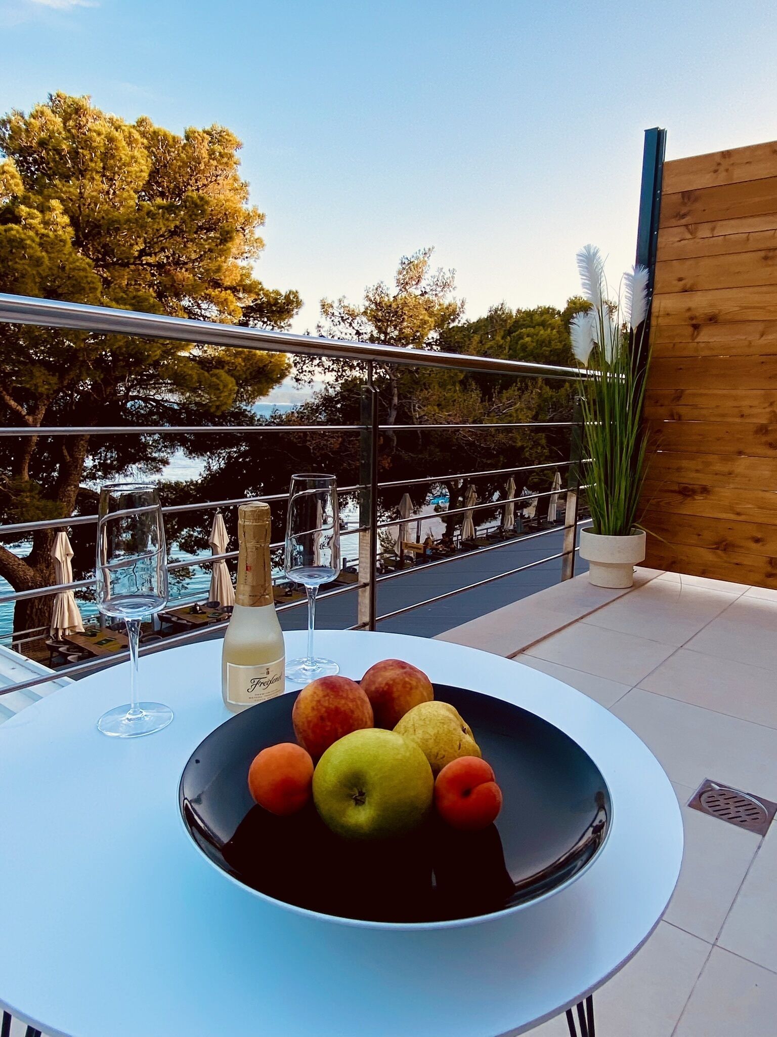 Una mesa con un cuenco de fruta y una botella de champán en un balcón con vistas a una piscina.