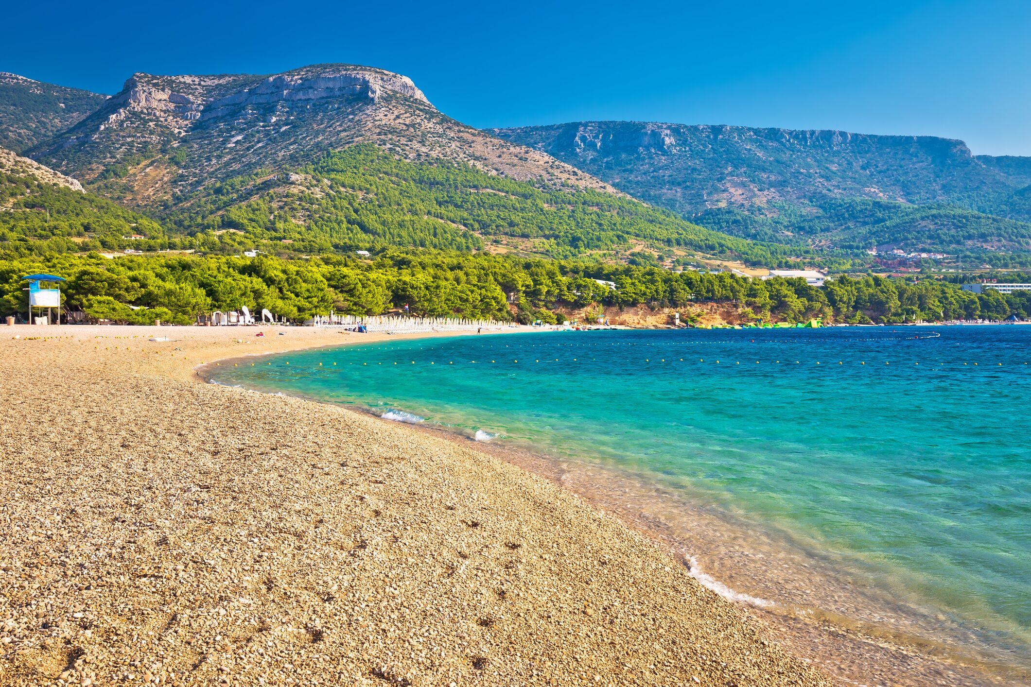 A pebble beach with clear turquoise waters, a mountain in the background, and a few buildings along the shore.