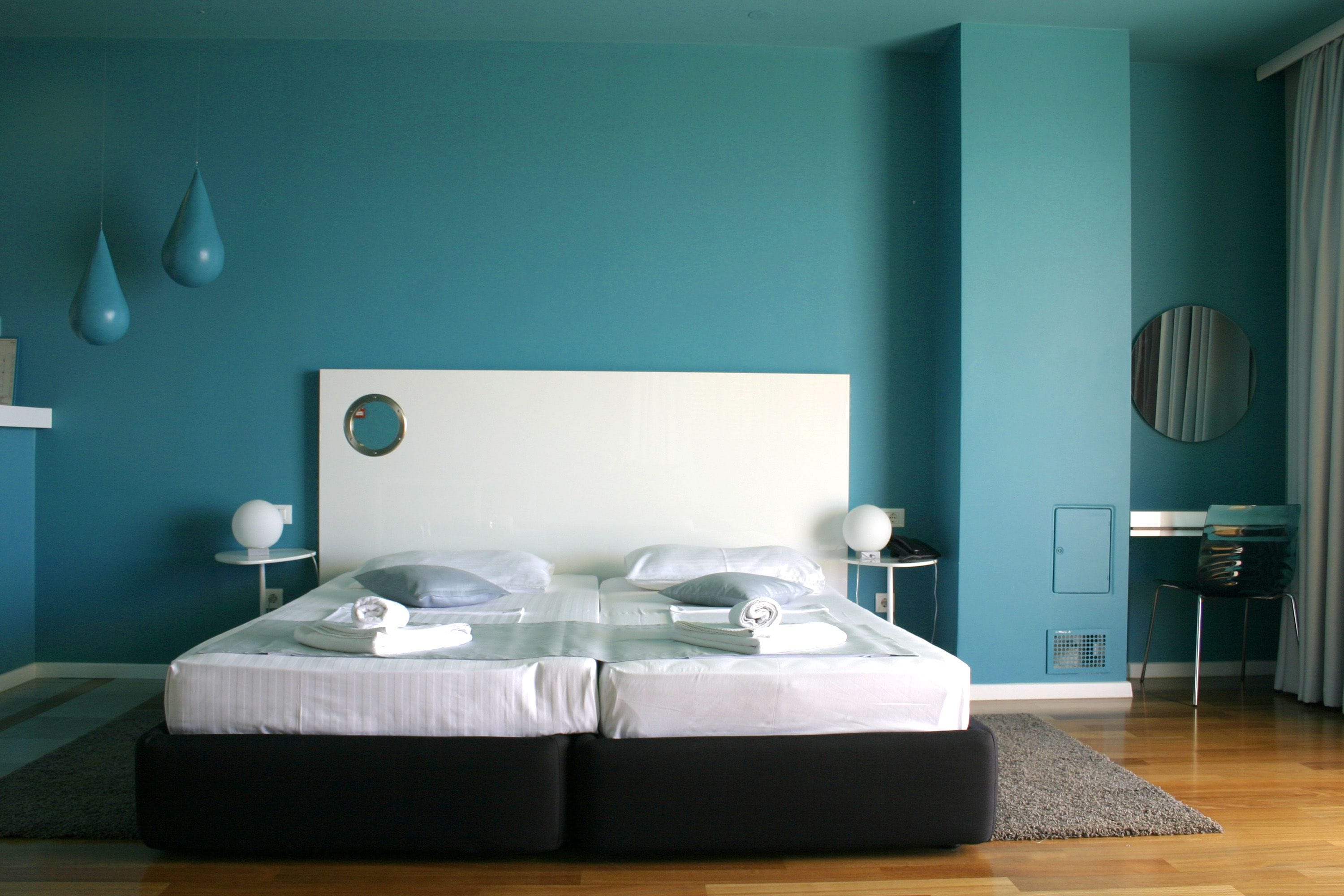 A modern bedroom with teal walls, a large bed with white bedding, two bedside tables with lamps, and a round mirror on the wall.