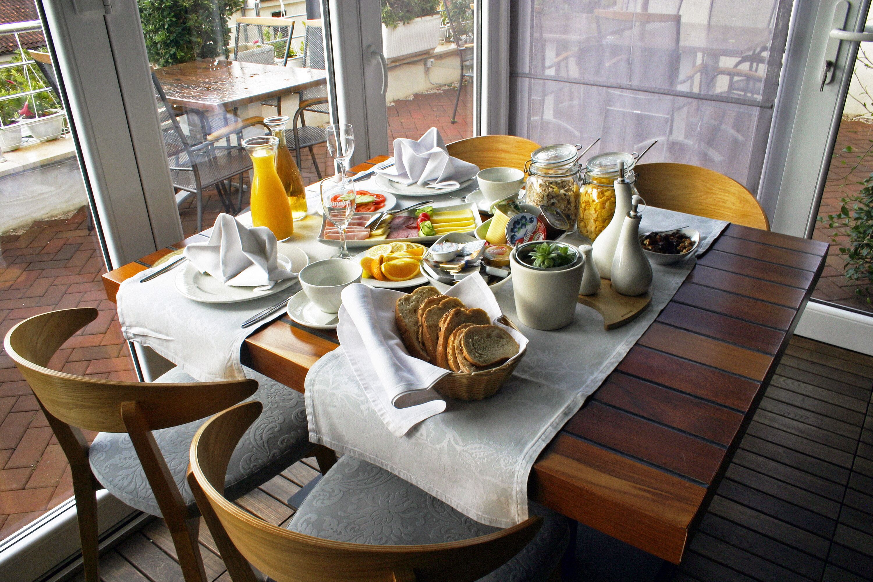 Une table de petit-déjeuner dressée avec divers aliments et boissons, entourée de chaises et offrant une vue sur l’extérieur.