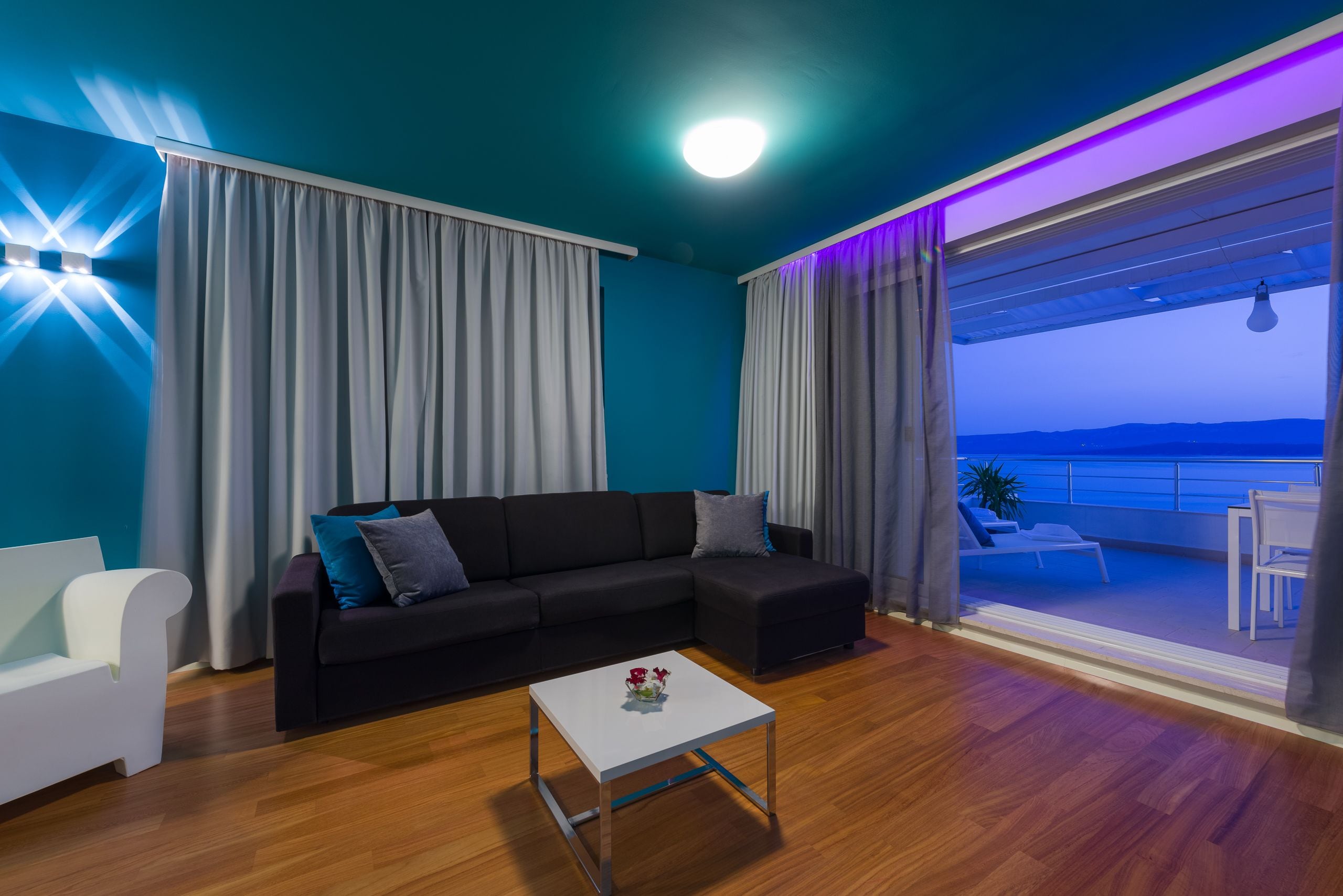A modern living room with a black sectional sofa, a white coffee table, and a view of a balcony with outdoor seating.