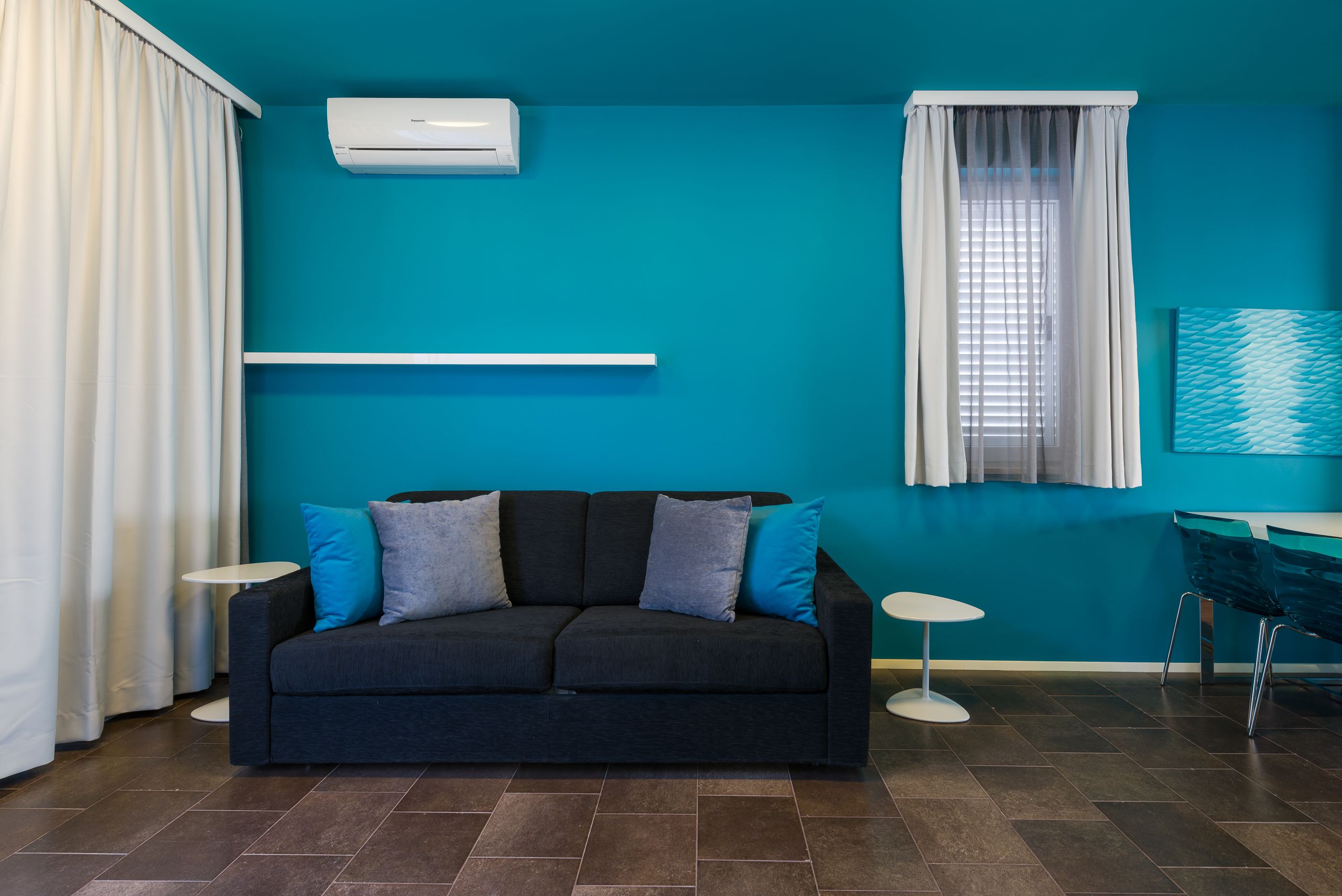 Un salon moderne avec un canapé foncé, deux tables d’appoint, une fenêtre avec des stores et un mur d’accent de couleur turquoise.