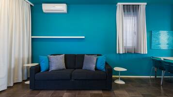 Un salon moderne avec un canapé foncé, deux tables d’appoint, une fenêtre avec des stores et un mur d’accent de couleur turquoise.