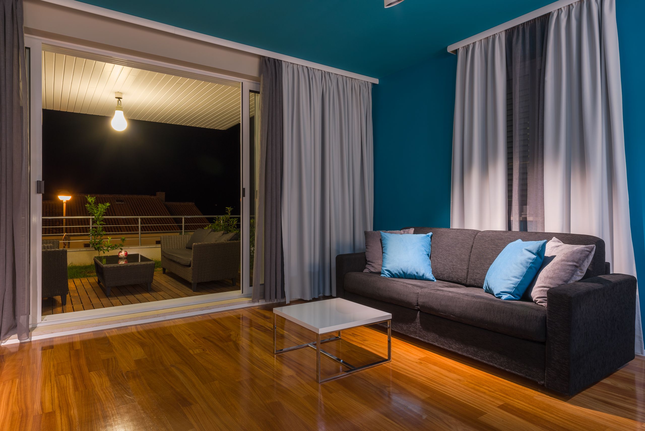 Uma sala de estar moderna com um sofá cinza escuro, uma mesa de centro branca e uma porta de vidro deslizante que dá para uma área externa com um pátio e um abajur.