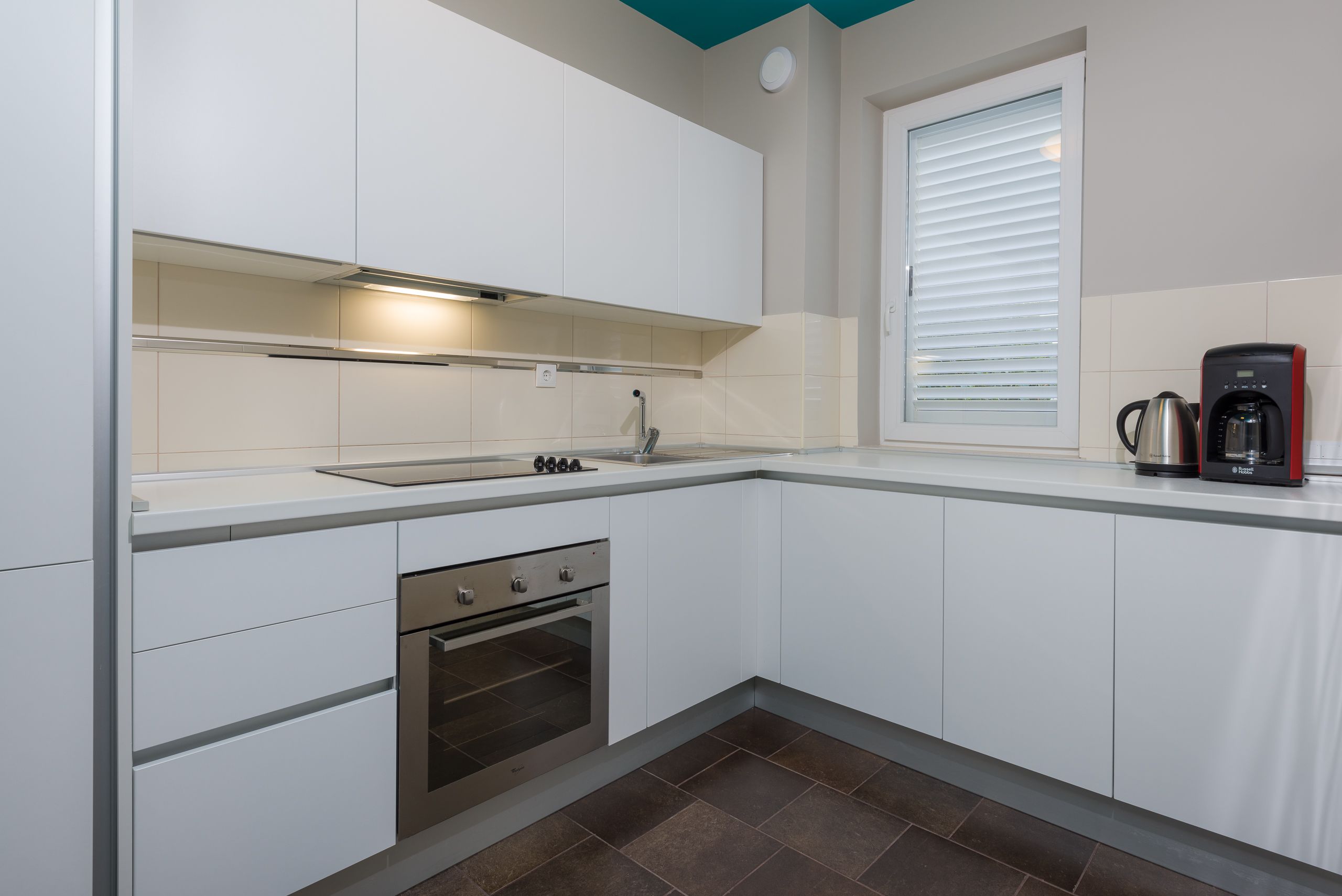 A modern kitchen with white cabinetry, a stainless steel oven, a sink, and a window with blinds.