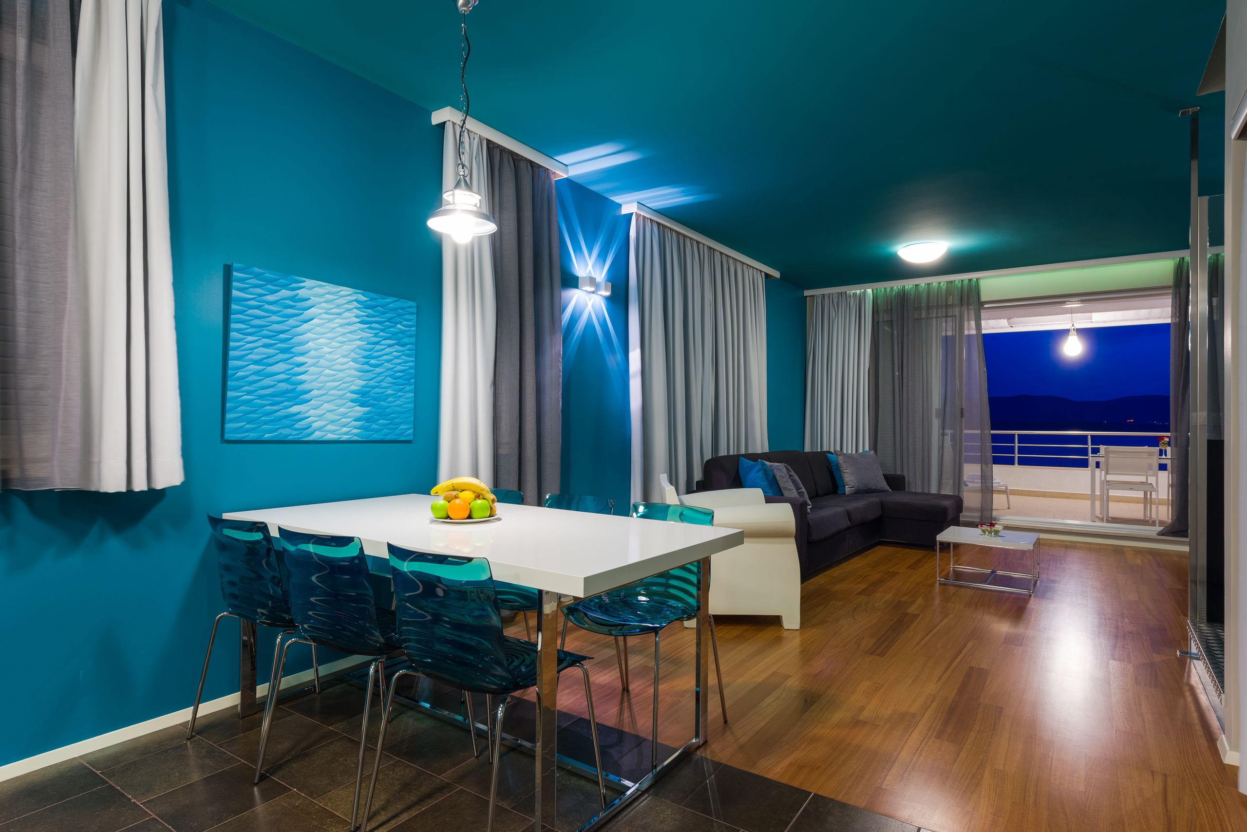 A modern living room with a dining area, featuring a white table and blue chairs, a sofa, and a balcony with a view.