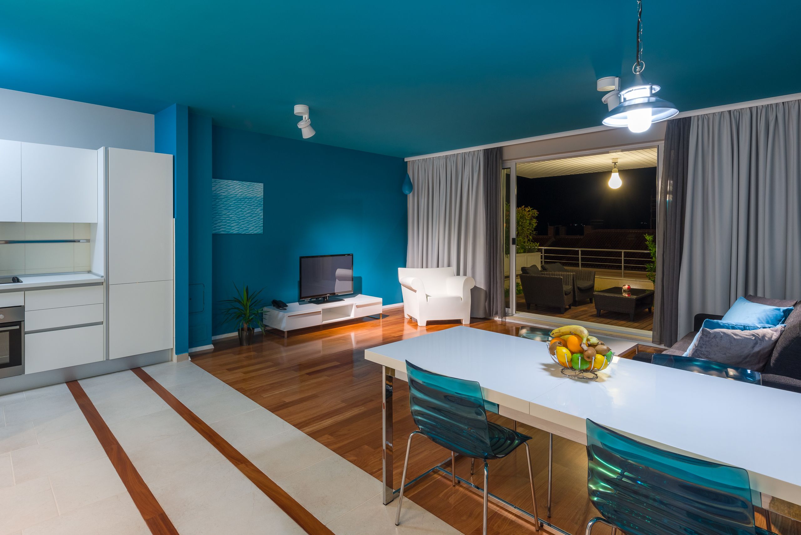 A modern kitchen with white cabinets, a dining area with a white table and blue chairs, a TV on a white stand, and a balcony with grey curtains.