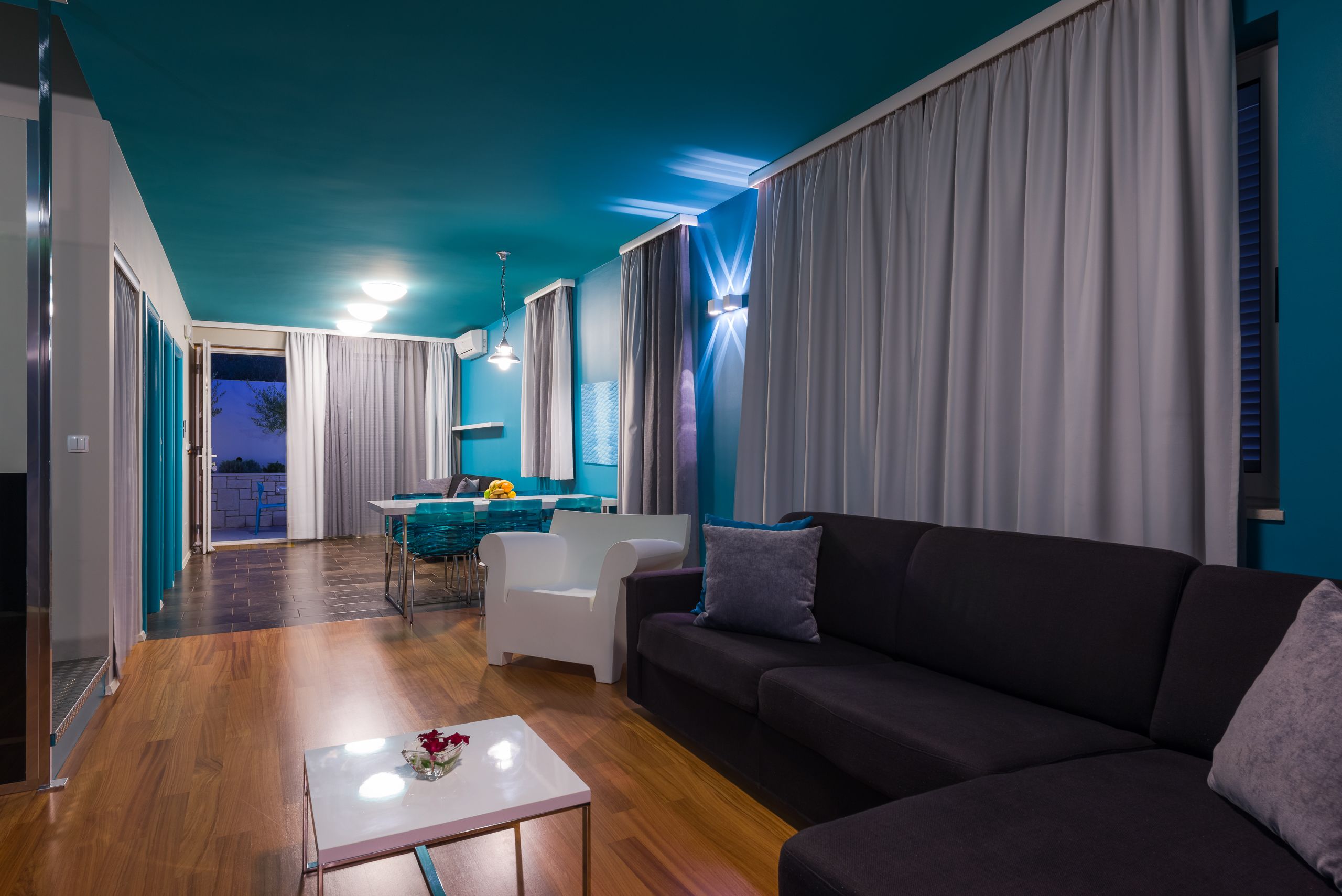 A modern living room with a dark sofa, a white coffee table, and a dining area with a blue table and chairs.