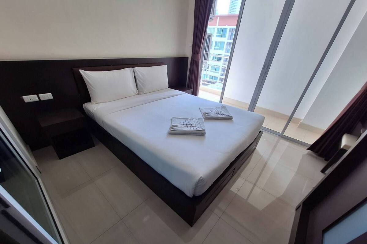 A modern hotel room with a large bed, a nightstand, a window with curtains, and a view of a building outside.