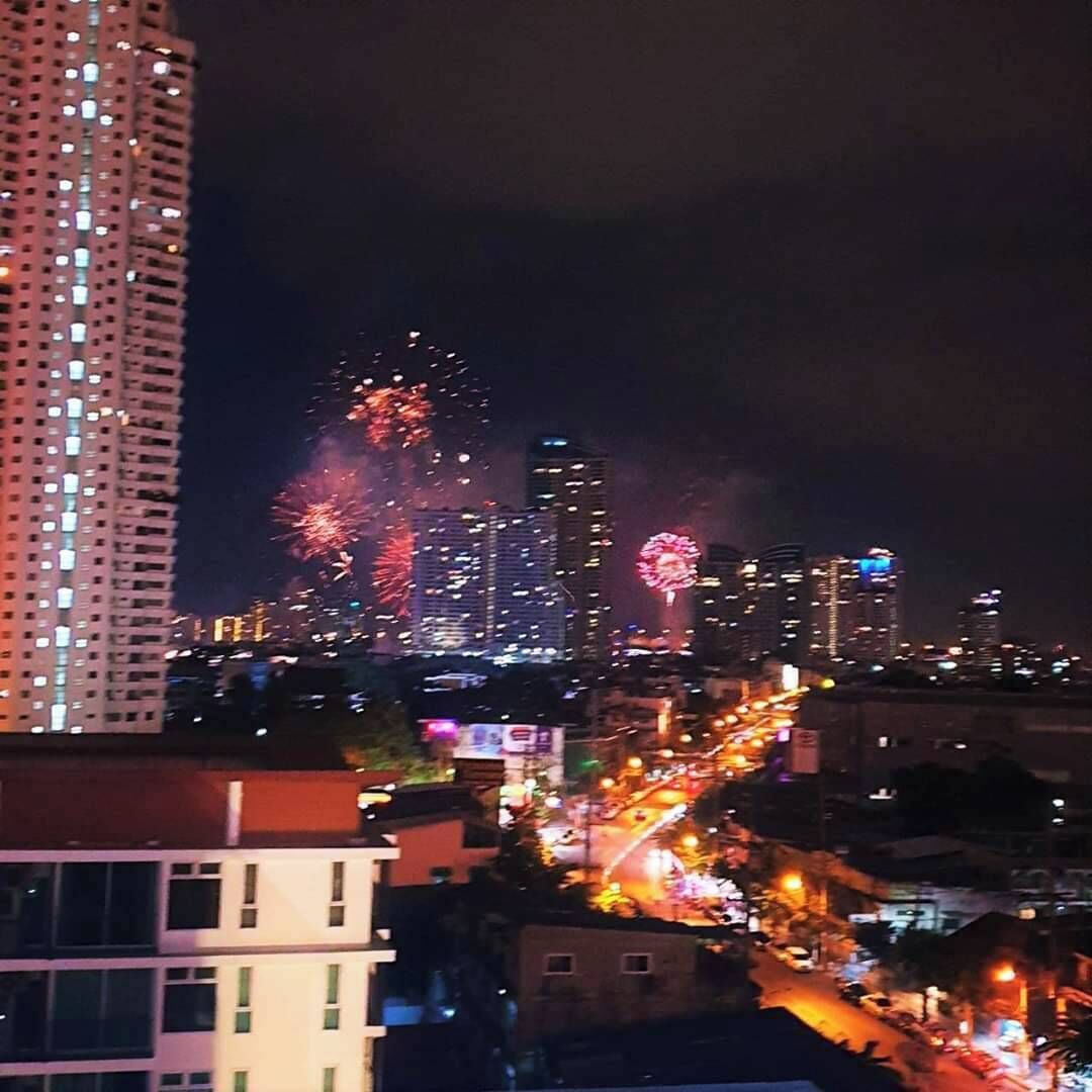 A nighttime cityscape with illuminated buildings and fireworks in the distance.