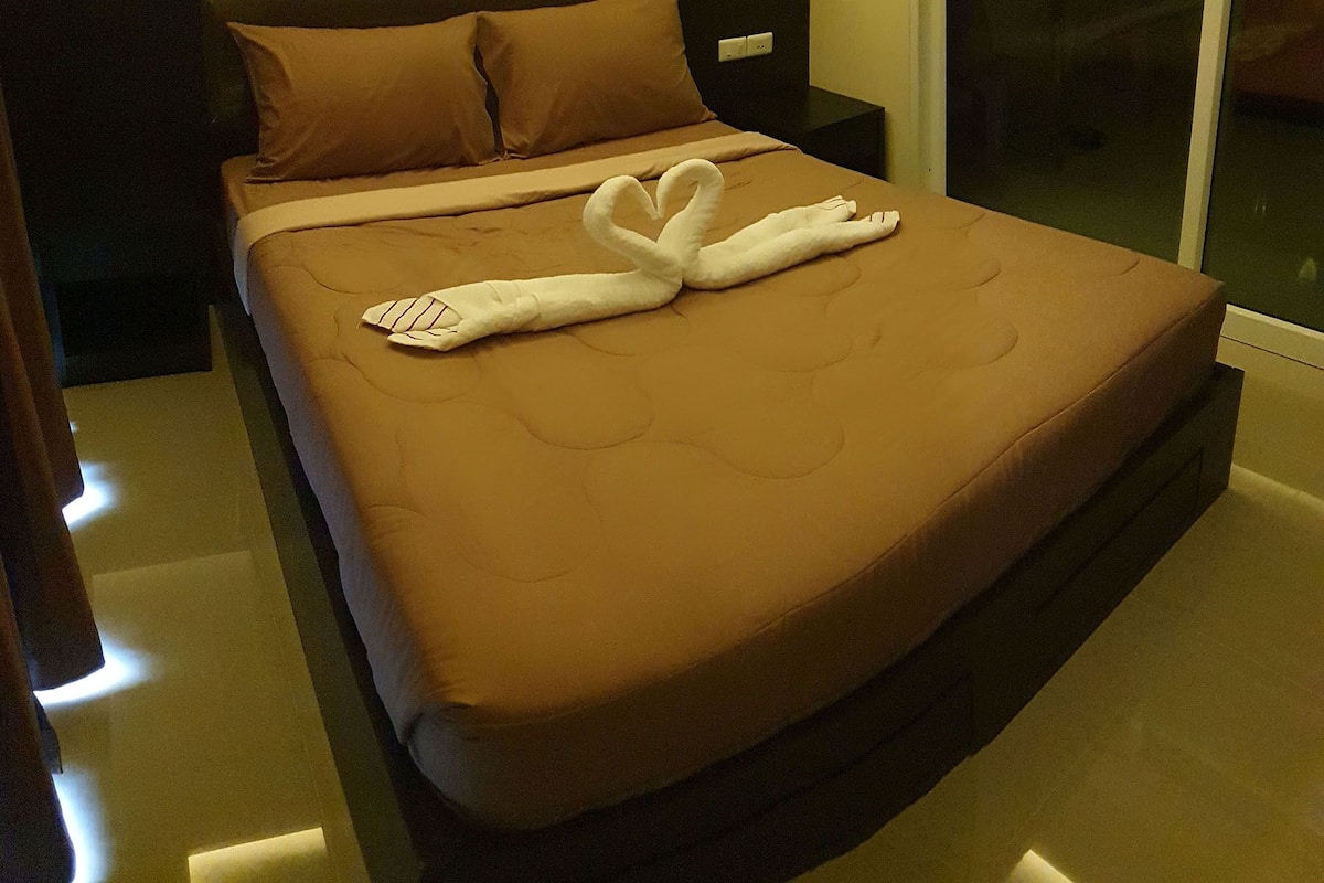 A neatly made bed with a brown mattress and pillows, a bedside table with a lamp, and a window with a view.