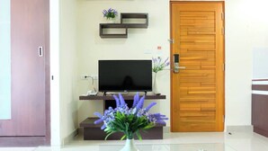 A modern living room with a flat-screen TV on a wooden stand, a wooden door, and a vase with purple flowers on a glass table. - The Cosy River (Bangkok)