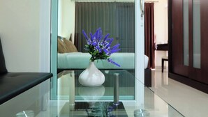 A modern hotel room with a glass partition, a white vase with purple flowers on a glass table, a bed with a white headboard, and a wooden wardrobe. - The Cosy River (Bangkok)