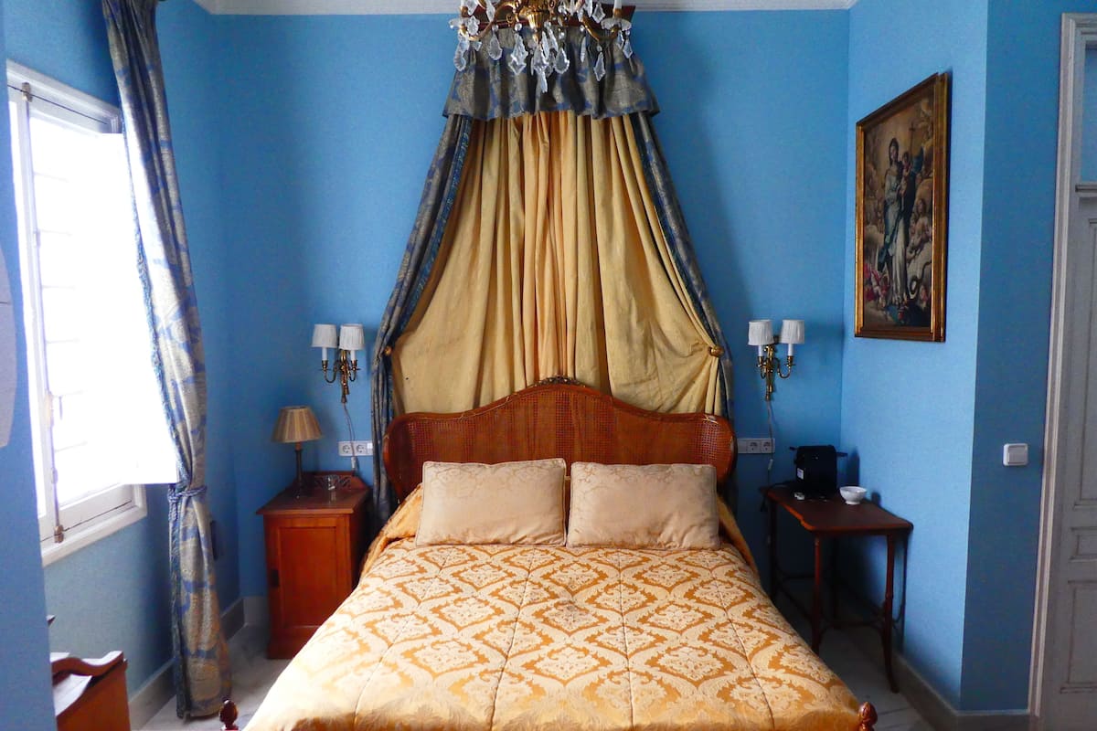 A bedroom with a large bed, a canopy, two bedside tables with lamps, a nightstand, a painting on the wall, and a window with curtains.