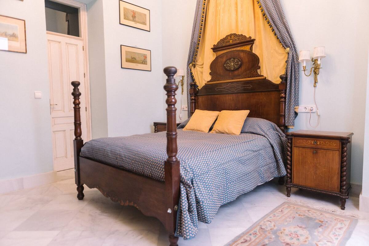 A four-poster bed with a canopy, a nightstand, a wall-mounted lamp, and framed pictures on the wall.