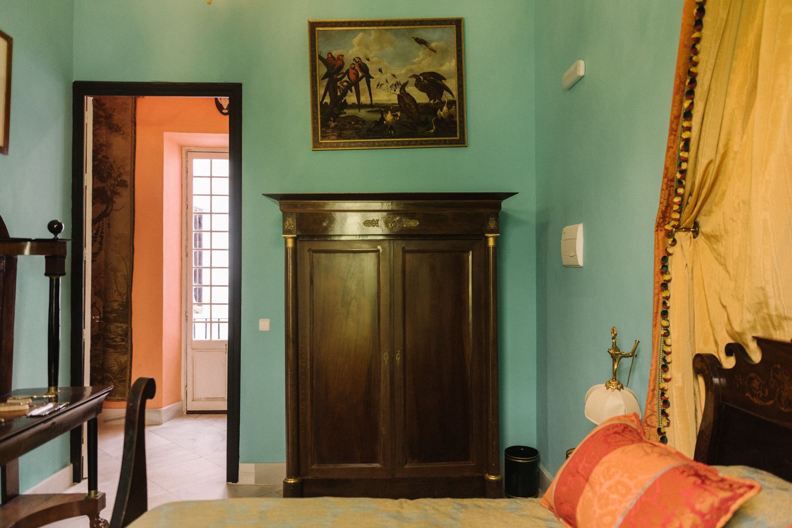 A room with teal walls, a wooden wardrobe, a bed with a red and orange pillow, and a painting above the wardrobe.