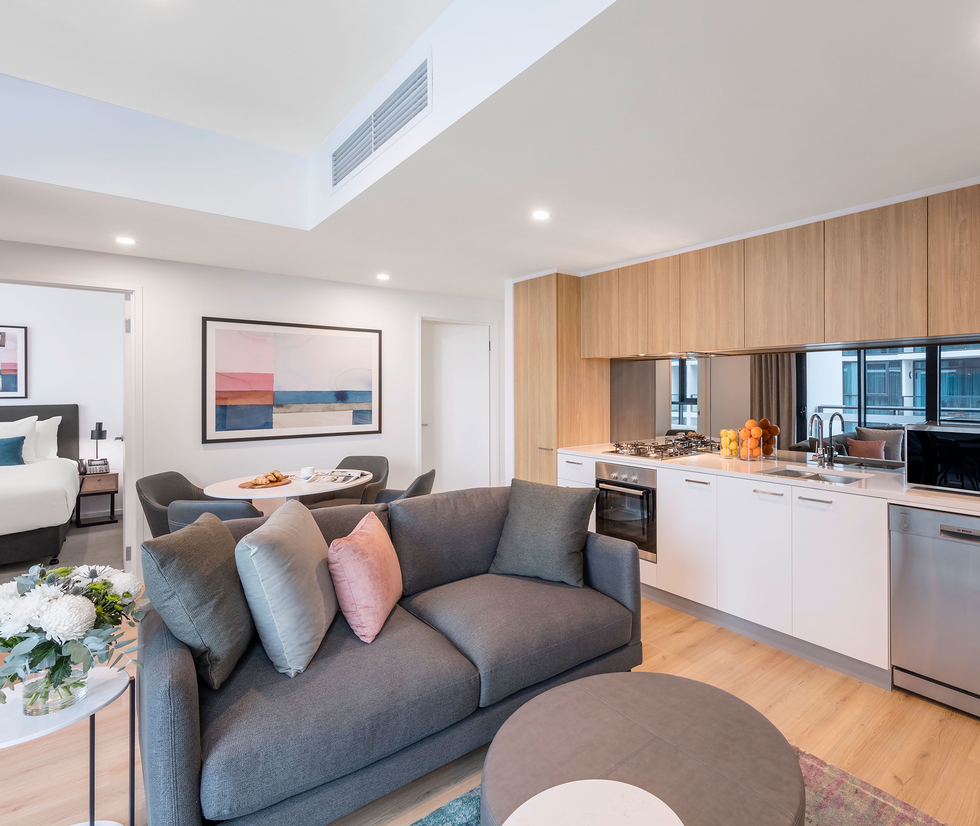A modern living room with a grey sofa, a dining table with chairs, and a kitchen area with white cabinets and a stove.