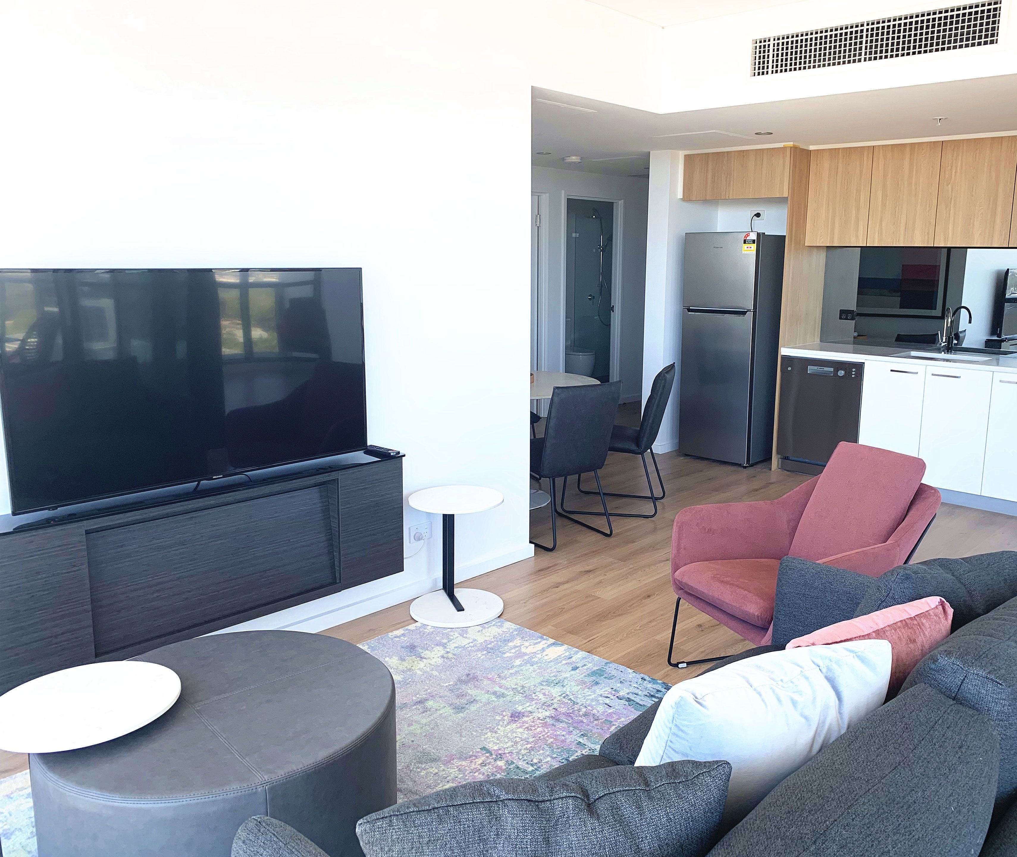 A modern living room with a flat-screen TV, grey sofas, a small round table, and a kitchen area with wooden cabinets and a stainless steel refrigerator.
