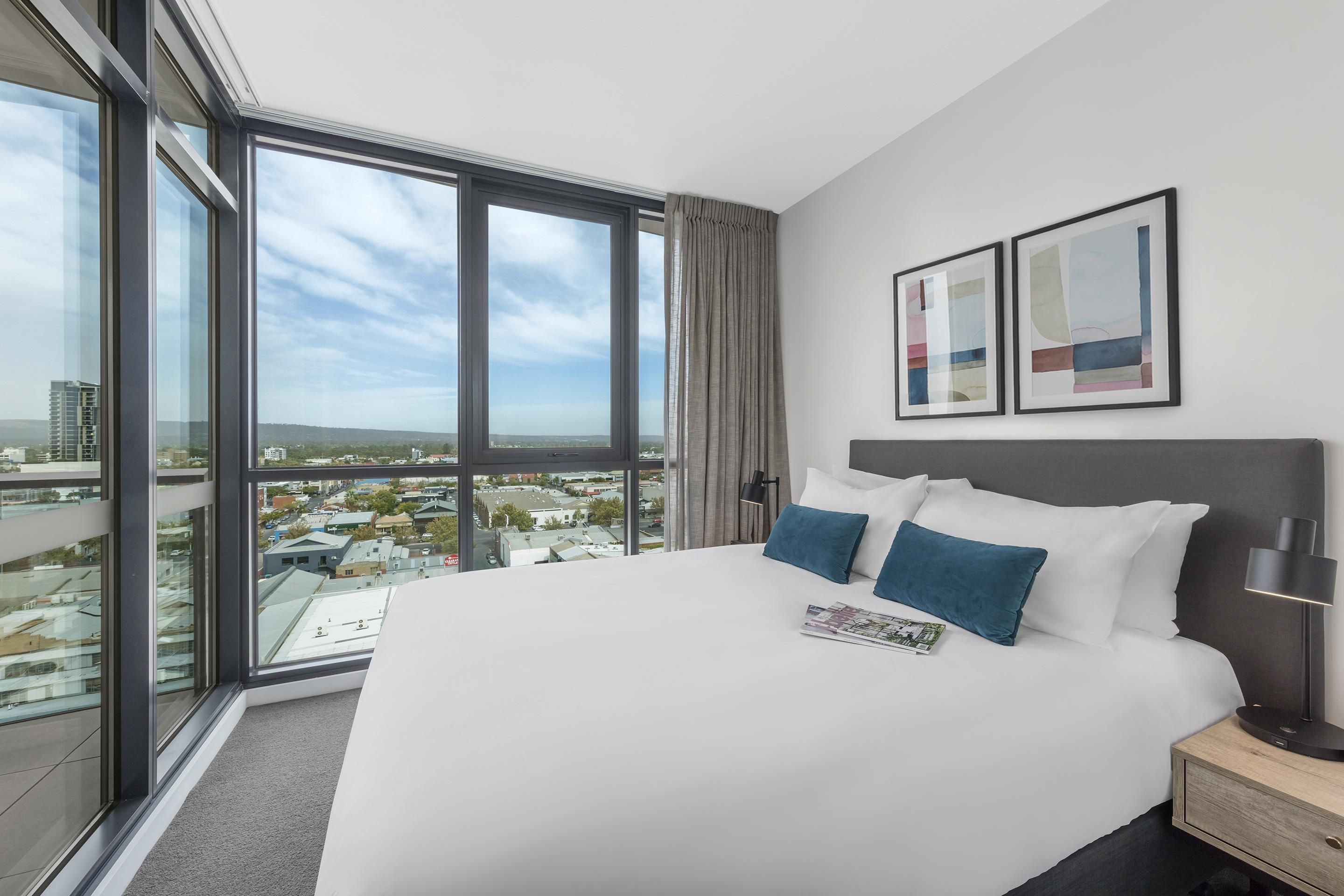 Ein Hotelzimmer mit einem großen Bett, zwei Kissen, einem Nachttisch, einer Lampe und Blick auf die Stadt durch große Fenster.