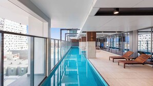 A modern indoor swimming pool with clear glass walls, wooden lounge chairs, and a view of the cityscape.