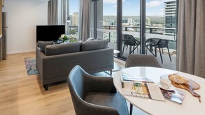 Una sala moderna con un sofá gris, un televisor de pantalla plana y un comedor con una mesa preparada para dos personas. Amplias ventanas ofrecen vistas a la ciudad.