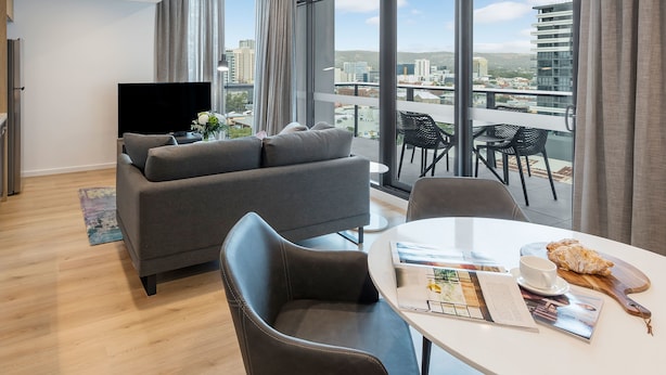 A modern living room with a grey sofa, a flat-screen TV, and a dining area with a table set for two. Large windows offer a city view.